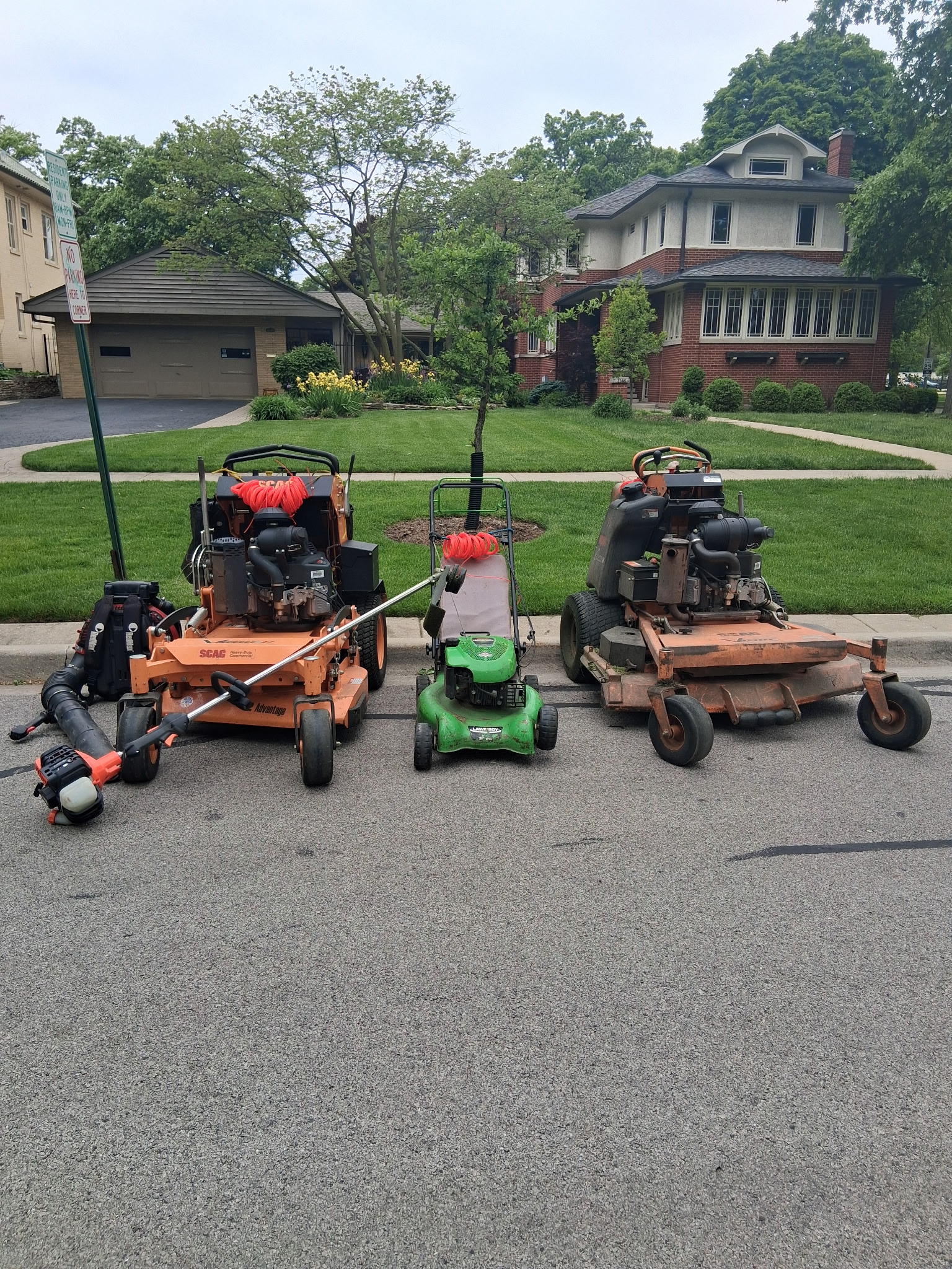 The full AJ Landscaping equipment lineup on the street: two SCAG commercial mowers, Lawn-Boy push mower, backpack blower, and string trimmer